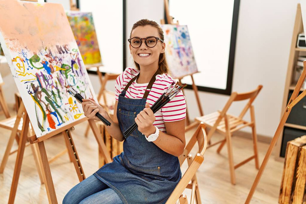 Νεαρή γυναίκα χαμογελά με αυτοπεποίθηση κρατώντας πινέλα στο στούντιο τέχνης - Φωτογραφία, εικόνα