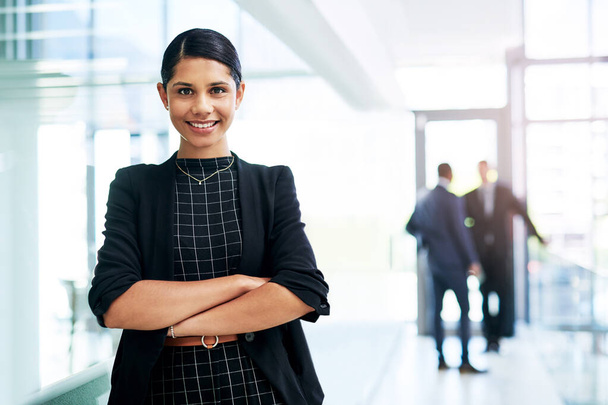 Im so glad I didnt doubt myself. Cropped portrait of an attractive young businesswoman standing with her arms folded in an office with her colleagues in the background - Photo, Image