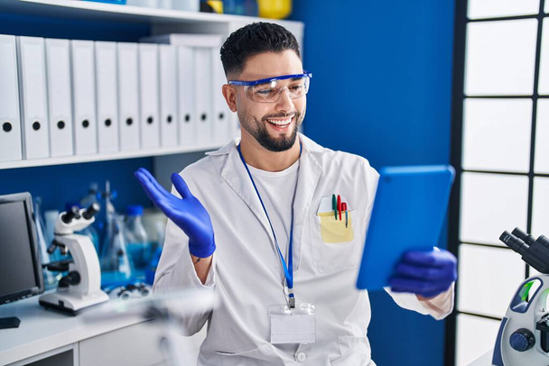 Mladý pohledný muž pracující v laboratoři vědec dělá on-line volání slaví úspěch se šťastným úsměvem a vítězný výraz se zvednutou rukou  - Fotografie, Obrázek