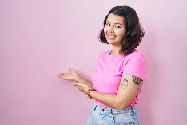 Young hispanic woman standing over pink background inviting to enter smiling natural with open hand  - Photo, Image