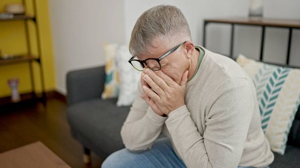 Hombre de pelo gris de mediana edad estresado sentado en el sofá llorando en casa - Foto, imagen