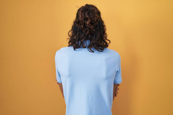 Junger hispanischer Mann steht auf gelbem Hintergrund und blickt rückwärts mit verschränkten Armen weg  - Foto, Bild