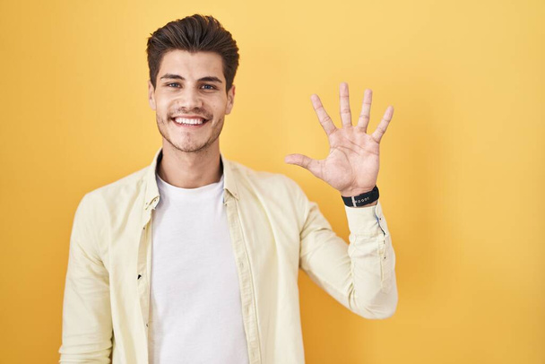 Sarı arka planda duran İspanyol genç adam beş numarayı gösterip işaret ederken kendinden emin ve mutlu gülümsüyor..  - Fotoğraf, Görsel