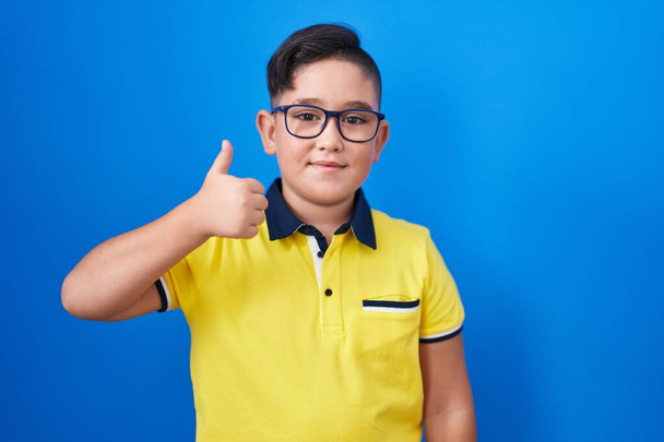 Young hispanic kid standing over blue background smiling happy and positive, thumb up doing excellent and approval sign  - Fotó, kép