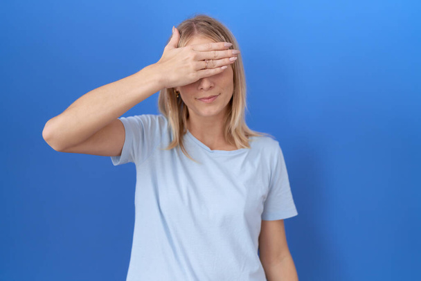 Młoda biała kobieta w luźnej niebieskiej koszulce zakrywającej oczy dłonią, wyglądająca poważnie i smutno. Koncepcja niewidoczności, ukrywania i odrzucania  - Zdjęcie, obraz