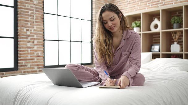 Junge schöne hispanische Frau mit Laptop schreiben auf Notizbuch im Schlafzimmer - Foto, Bild