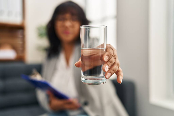 Молодая красивая латинская женщина-психолог улыбается уверенно предлагая стакан воды в психологической клинике - Фото, изображение