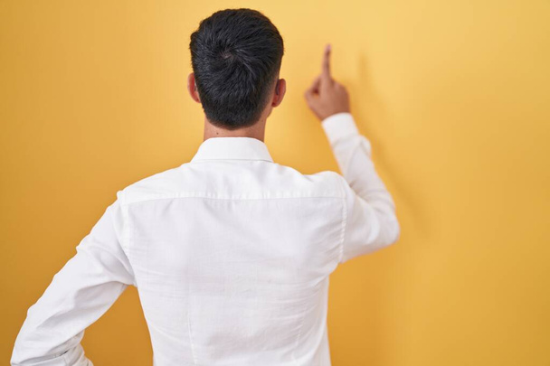 Young hispanic man standing over yellow background posing backwards pointing ahead with finger hand  - 写真・画像