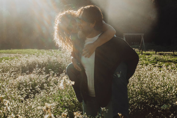 Szerelem pár élvezi naplemente és szabadtéri szabadidős tevékenység együtt a természetben. Férfi cipeli a nőt a hátán a romantikus festői réten fehér virágos virágokkal. vándor úti cél napja - Fotó, kép