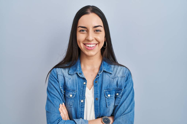 Latijns-Amerikaanse vrouw die over een blauwe achtergrond staat... vrolijk gezicht glimlachend met gekruiste armen naar de camera kijkend. positieve persoon.  - Foto, afbeelding