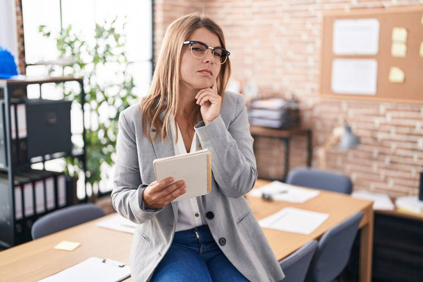 Young hispanic woman working at the office wearing glasses with hand on chin thinking about question, pensive expression. smiling with thoughtful face. doubt concept.  - Фото, изображение