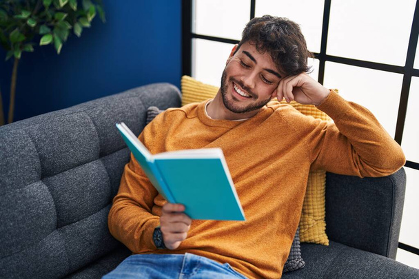 Νεαρός Ισπανός διαβάζει βιβλίο καθισμένος στον καναπέ στο σπίτι - Φωτογραφία, εικόνα