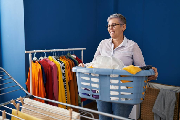 Kobieta w średnim wieku uśmiechnięta pewna siebie trzymając kosz z ubraniami w pralni - Zdjęcie, obraz
