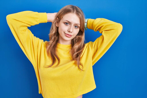 Young caucasian woman standing over blue background relaxing and stretching, arms and hands behind head and neck smiling happy  - Foto, afbeelding