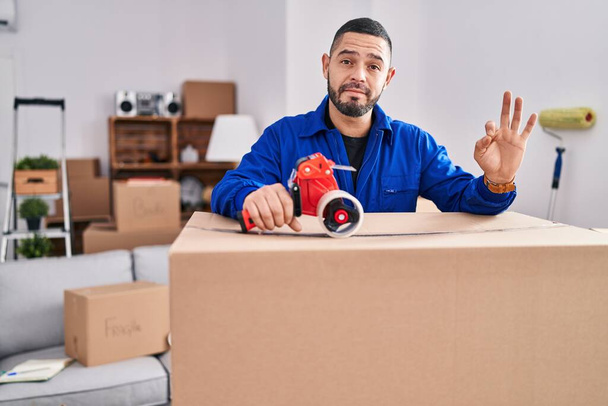 Hispanic man working on moving holding packing tape doing ok sign with fingers, smiling friendly gesturing excellent symbol  - Photo, Image