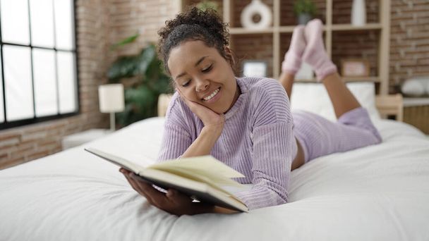 Mujer afroamericana leyendo libro acostada en la cama en el dormitorio - Foto, imagen