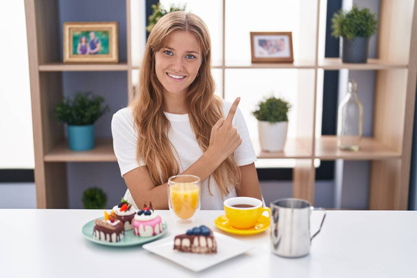 Молодая белая женщина ест выпечку на завтрак весёлая с улыбкой на лице, указывающей рукой и пальцем в сторону, со счастливым и естественным выражением лица  - Фото, изображение
