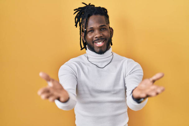Homme africain avec dreadlocks portant pull col roulé sur fond jaune souriant joyeux offrant mains donnant assistance et acceptation.  - Photo, image