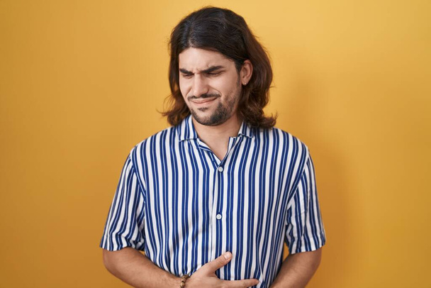 Hispanic man with long hair standing over yellow background with hand on stomach because nausea, painful disease feeling unwell. ache concept.  - Photo, Image
