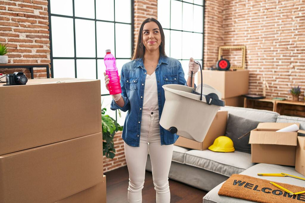 Latino nainen siirtymässä uuteen kotiin puhdistus lattia hymyillen sivuun ja tuijottaen pois ajattelu.  - Valokuva, kuva