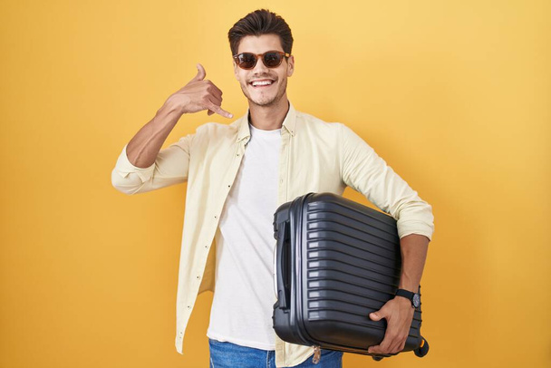 Yaz tatiline giderken bavul tutan genç İspanyol adam el ve parmaklarıyla telefonla konuşuyor gibi gülümsüyor. iletişim kavramları.  - Fotoğraf, Görsel
