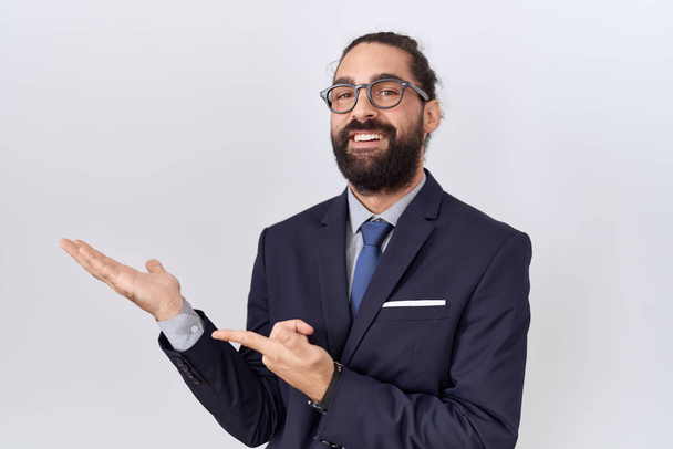 Latinomies, jolla on parta yllään puku ja solmio hämmästynyt ja hymyilee kameralle samalla, kun hän esittelee kädellään ja osoittaa sormella.  - Valokuva, kuva