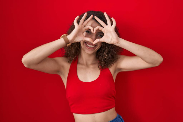 Hispanic woman with curly hair standing over red background doing heart shape with hand and fingers smiling looking through sign  - Фото, изображение