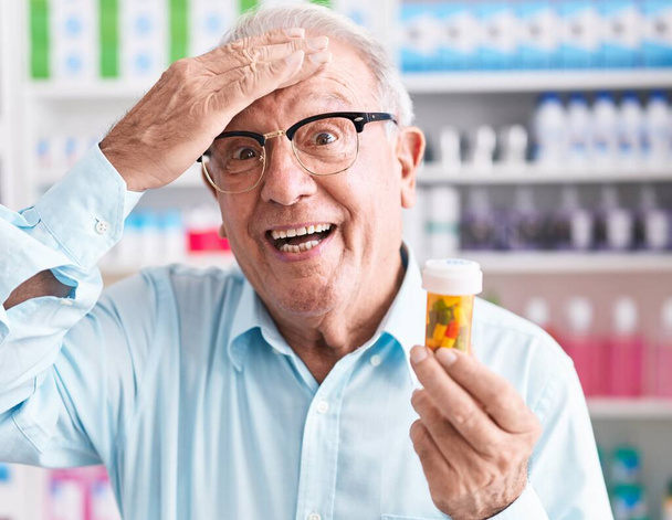 Hombre mayor con pelo gris sosteniendo pastillas en la farmacia estresado y frustrado con la mano en la cabeza, cara sorprendida y enojada  - Foto, imagen