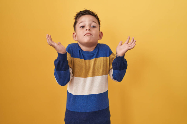 Μικρό ισπανόφωνο αγόρι στέκεται πάνω από κίτρινο φόντο φωνάζοντας και ουρλιάζοντας δυνατά στο πλάι με το χέρι στο στόμα. έννοια επικοινωνίας.  - Φωτογραφία, εικόνα