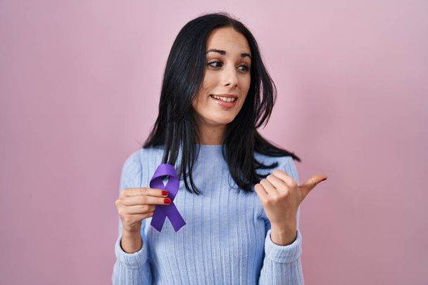Іспаномовна жінка тримає фіолетову стрічку усвідомлення, вказуючи великий палець на стороні посміхаючись щасливий відкритим ротом  - Фото, зображення