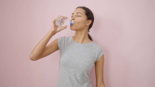 Молодая красивая латиноамериканка в спортивной одежде пьет воду на изолированном розовом фоне - Фото, изображение