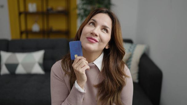 Joven mujer hispana hermosa sosteniendo tarjeta de crédito sentada en el sofá en casa - Foto, imagen