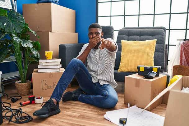 Hombre afroamericano sentado en el suelo en una casa nueva riéndose de ti, señalando con el dedo a la cámara con la mano sobre la boca, expresión de vergüenza  - Foto, Imagen