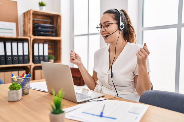 Jonge Spaanse vrouw draagt call center agent headset op kantoor erg blij en enthousiast doen winnaar gebaar met armen omhoog, glimlachen en schreeuwen om succes. viering concept.  - Foto, afbeelding