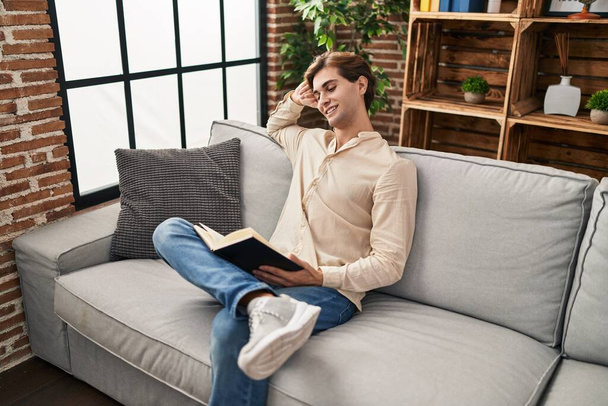 Young caucasian man reading book sitting on sofa at home - Valokuva, kuva
