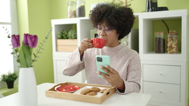 Giovane donna afroamericana che fa colazione utilizzando lo smartphone a casa - Foto, immagini
