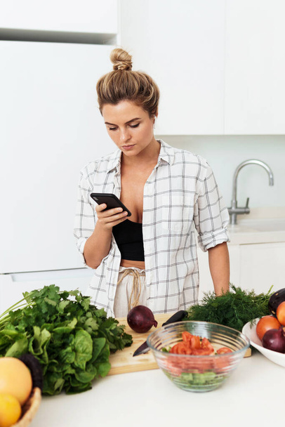 Young pretty woman with wireless earbuds is using smartphone during cooking in modern white kitchen - Foto, immagini