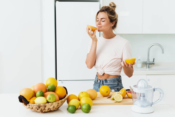Young beautiful woman sniffing fresh orange during citrus juice preparation at home - Foto, Imagem