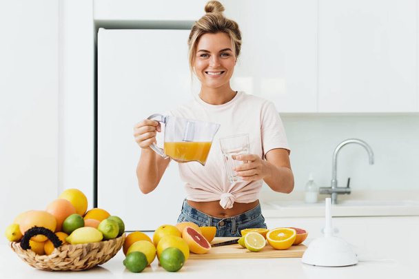 Young woman pouring freshly squeezed homemade orange juice into the glass in modern white kitchen - Foto, Bild