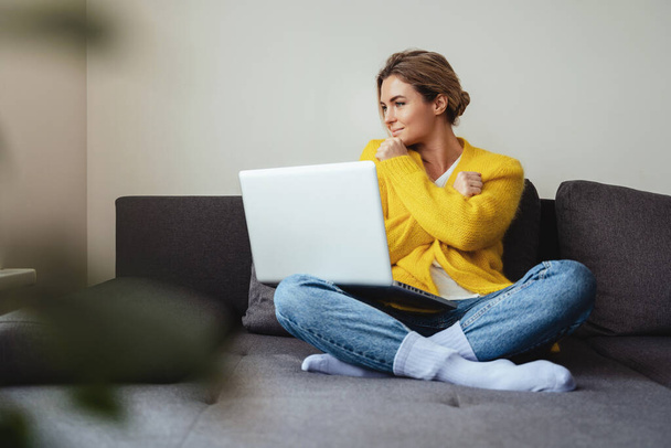 Young woman wearing yellow cardigan sitting on the sofa and using laptop computer - Foto, Imagem
