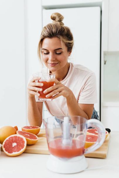 Young woman drinking freshly squeezed homemade grapefruit juice in white kitchen - Fotó, kép