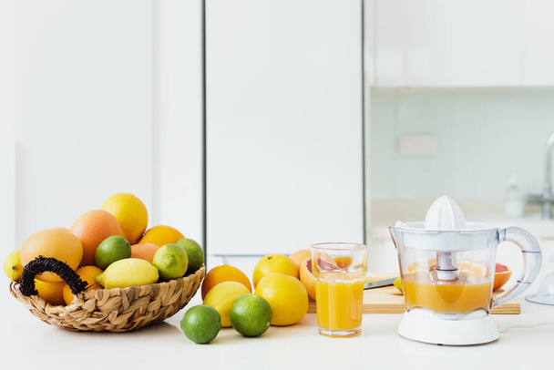 Glass of orange juice, citrus juicer and various citrus fruits on white kitchen table - Fotó, kép
