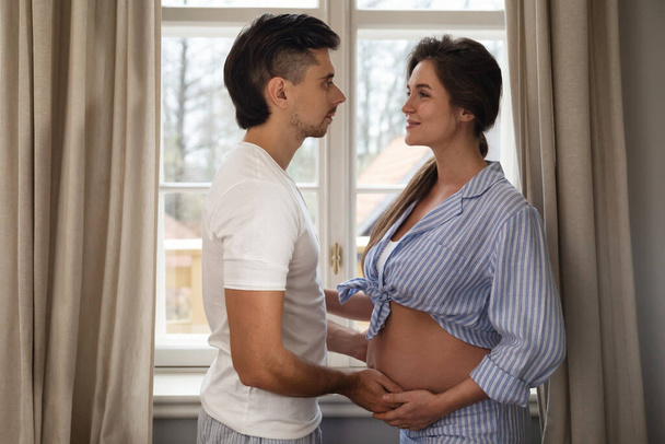Young sensual couple are waiting for a baby. Man and his pregnant wife in embrace beside the window.  - Foto, afbeelding