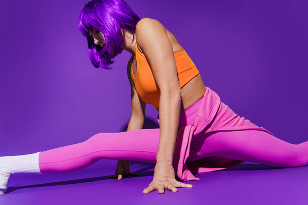Young woman wearing colorful sportswear doing splits against purple background - Photo, image