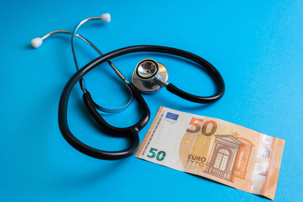 Closeup shot of a fifty euro banknote and a stethoscope on blue background. Concept of health care and community medicine price. - Foto, imagen
