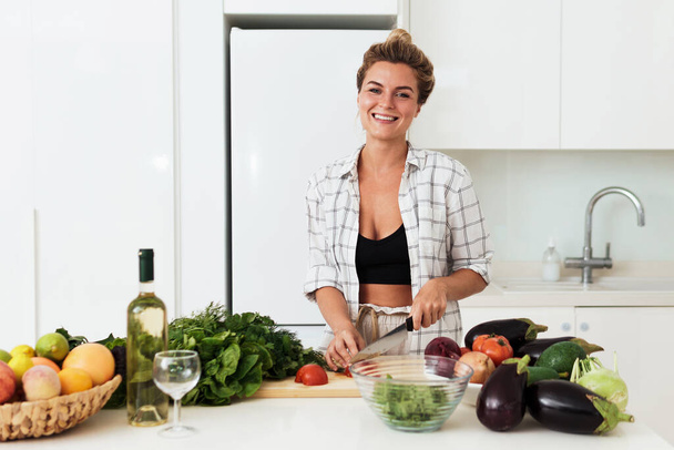 Young cheerful woman cooking healthy vegetarian dish at home in white kitchen - 写真・画像