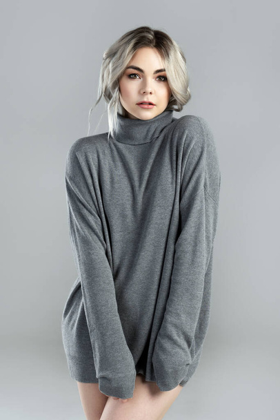 Portrait of young cute woman with blond hair wearing turtleneck jumper is posing on gray background in studio - Fotografie, Obrázek