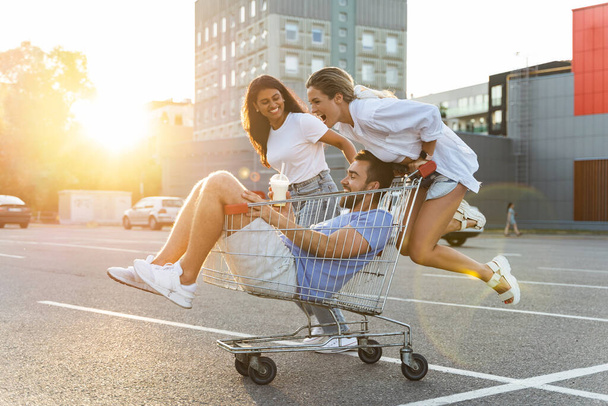 Three diverse friends having fun and riding shopping cart on parking lot near supermarket during summer evening - Zdjęcie, obraz