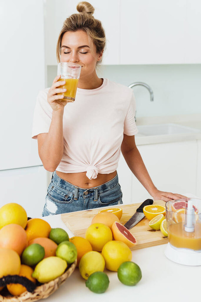 Young woman drinking freshly squeezed homemade orange juice in white kitchen - Foto, imagen