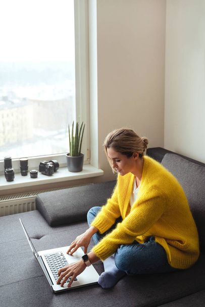 Mujer joven con cárdigan amarillo sentado en el sofá y el uso de ordenador portátil - Foto, Imagen
